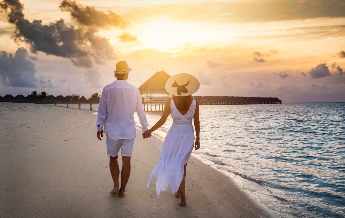 A beautiful traveler couple in white summer clothing walks along a tropical beach holding hands during sunset time